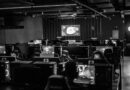 A black-and-white photograph of a gaming arena filled with rows of computer stations, monitors, and gaming chairs, with several players wearing headsets.