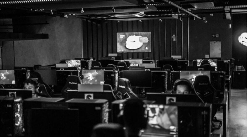 A black-and-white photograph of a gaming arena filled with rows of computer stations, monitors, and gaming chairs, with several players wearing headsets.