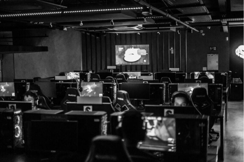 A black-and-white photograph of a gaming arena filled with rows of computer stations, monitors, and gaming chairs, with several players wearing headsets.